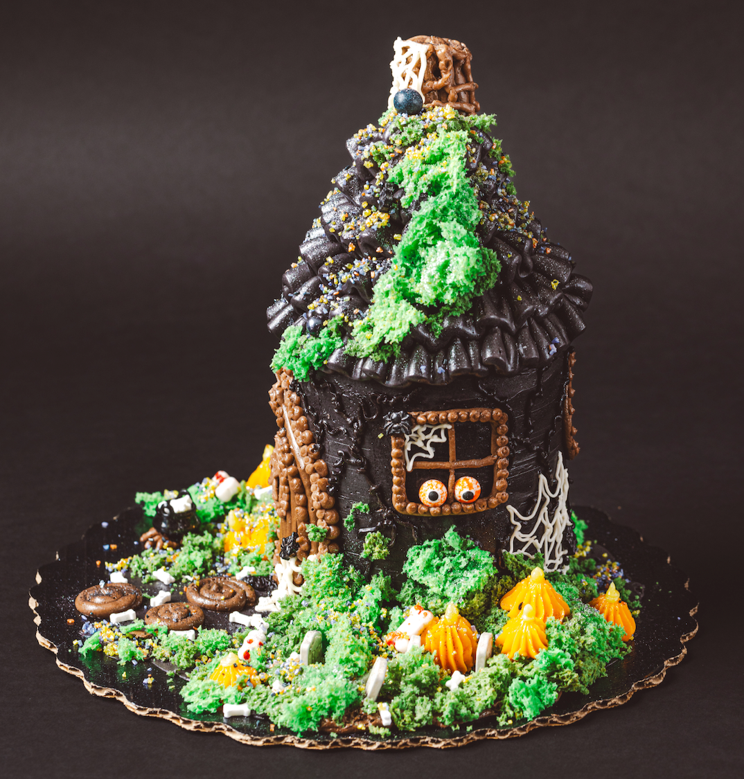 Decorative cake shaped like a haunted house with greenery and small figures on a dark background