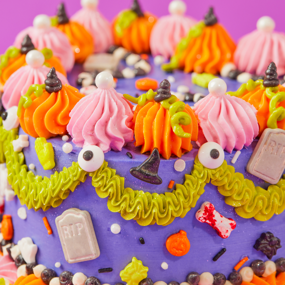 
                  
                    Colorful Halloween-themed cake with purple base and various decorations on a purple background
                  
                