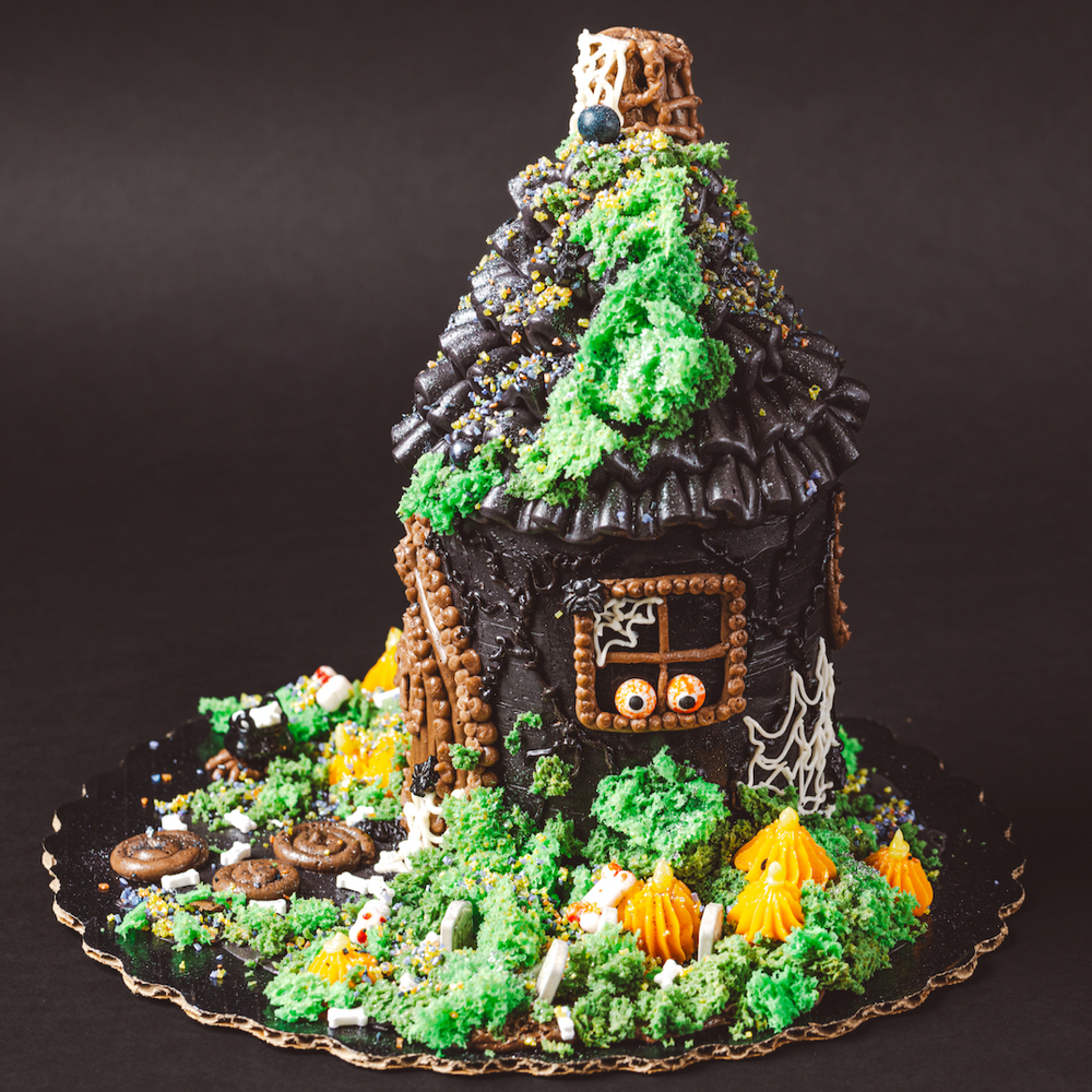 Decorative cake shaped like a haunted house with greenery and small figures on a dark background